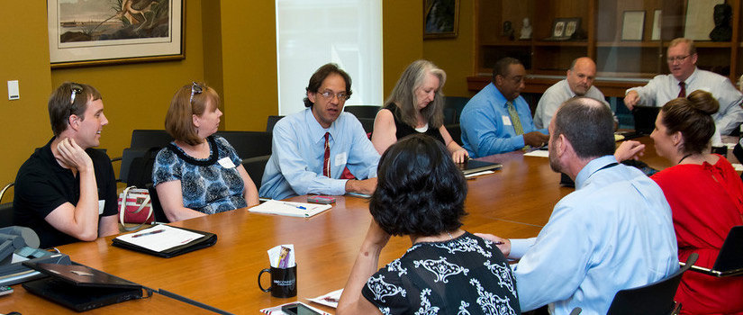 University committee members sitting around a conference table and listening to one person speak to the group. 