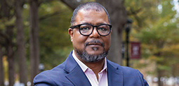 A man with glasses on standing outside in a gray suite. 