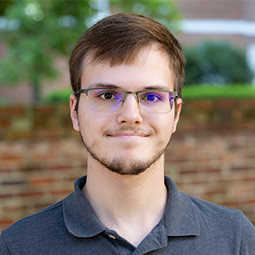 photo of a man with short brown hair and glasses wearing a gray shirt while standing outside