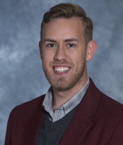 Christopher wears a garney blazer and a gray sweater and smiles at the camera