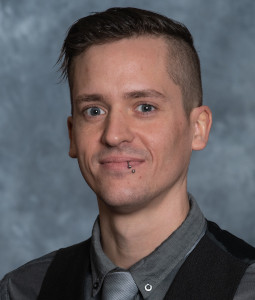 Dustin wears a gray oxford and black vest and smiles at the camera