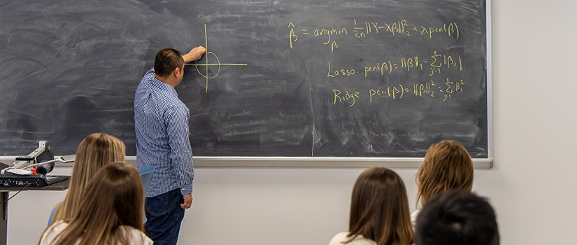 professor at a black board writing formulas while students watch and take notes