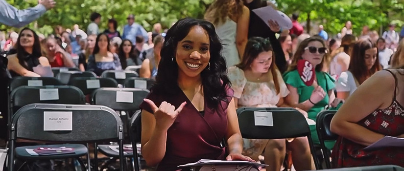 A young black woman sitting down smiling as she has a graduation cap on her lap.