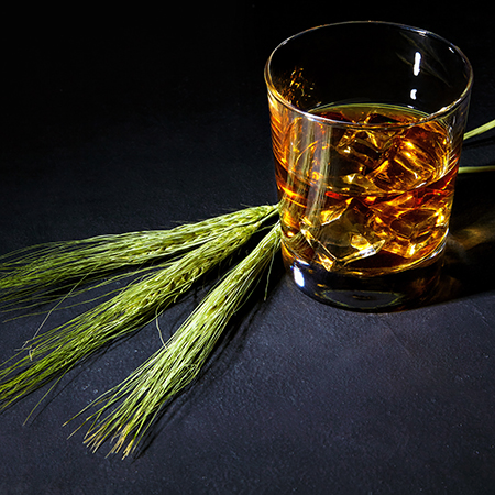 Glass of whiskey with ice in a glass cup surrounded by ears of wheat on a black stone table.