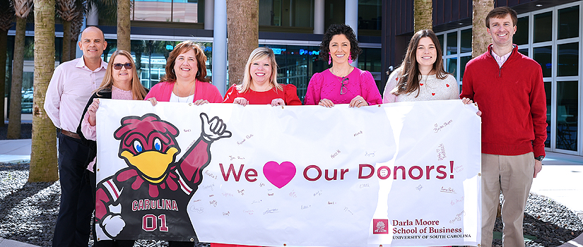 Development staff members standing in the Moore School courtyard, holding a banner with Cocky on it and the text: We love our donors!