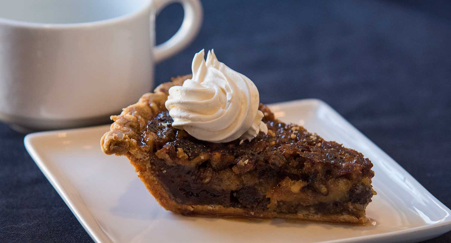 Slice of pie on a white plate with a cup of coffee sit on a blue table cloth.