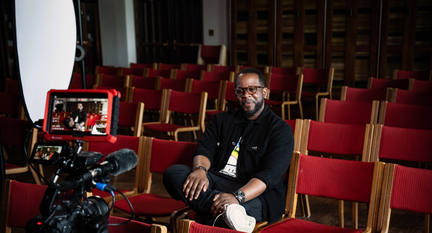 Camera films Jabari Evans while he sits in a room full of red chairs.