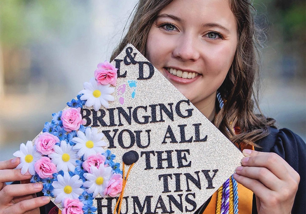 Graduate holding a commencement cap reading 'Bringing you all the tiny humans.'